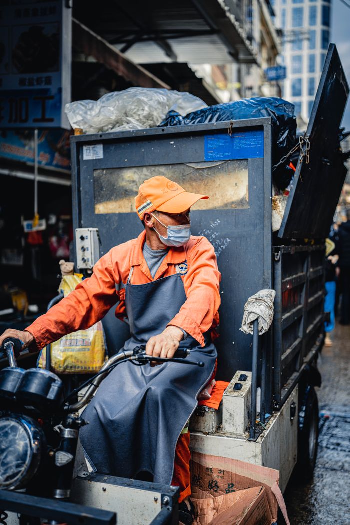 Crafting Captivating Headlines: Your awesome post title goes here Male worker wearing uniform and protective mask driving transport with rubbish on sidewalk of local street in city with buildings